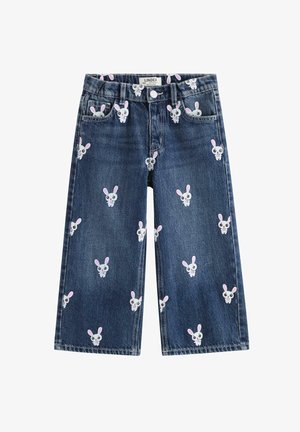 Smilende barn med krøllet hår iført en hvid t-shirt, blå jeans med kaninprint og hvide sneakers, stående mod en ensfarvet baggrund.