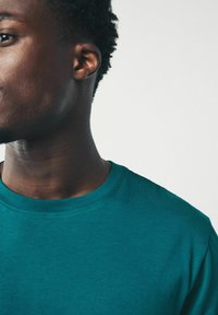 Túrkiz színű, kerek nyakú, puha anyagból készült póló. A közeli felvétel sima textúrát és finom varrási részleteket mutat be a nyakrészen és az ujjakon.