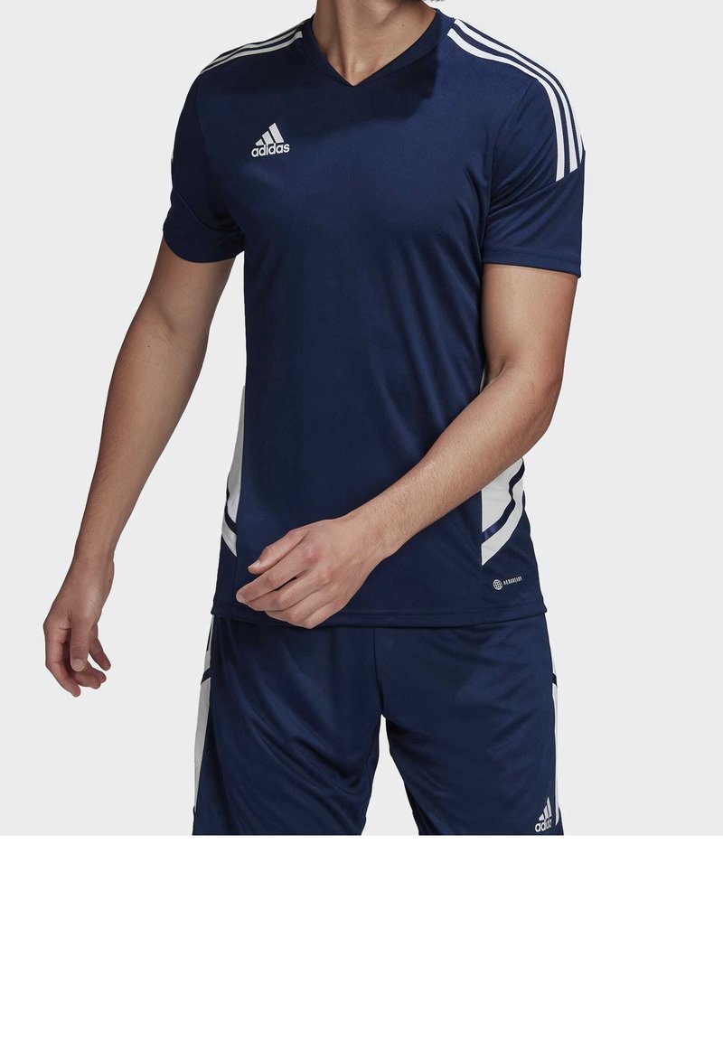 Homme portant un maillot et un short de sport Adidas bleu marine avec des bandes blanches et des logos, debout devant un fond uni.