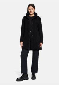 Zwarte wolmix jas met capuchon, voorzien van vier knopen aan de voorkant, lange mouwen en een gestructureerde afwerking. Gecombineerd met donkere broeken en chunky laarzen.