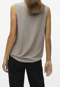 Top senza maniche di un leggero color taupe con finitura texturizzata, vestibilità morbida e orlo arrotondato; abbinato a pantaloni neri con dettagli sulle tasche posteriori.