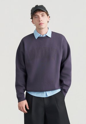 Tummanvioletti ylisuurikokoinen collegepaita, jossa on teksturoitu logo, kerrospukeutuneena vaaleansinisen kauluspuseron kanssa, yhdistettynä mustiin housuihin ja lippalakkiin.