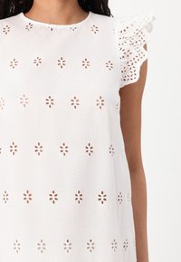 Witte katoenen jurk met geborduurde bloemenuitsnijdingen, gerimpelde kanten mouwen en een ronde halslijn, die een gelaagde, lichte uitstraling geeft.