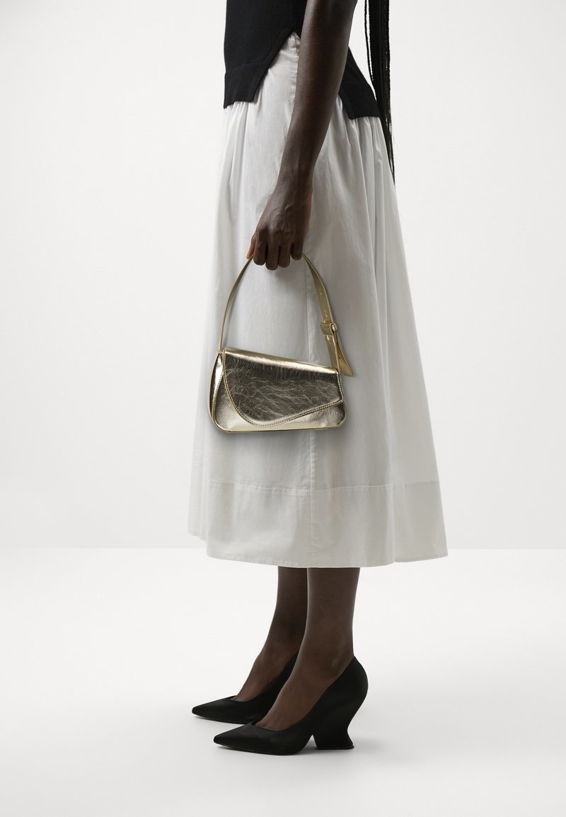 Femme portant une jupe midi blanche et des chaussures compensées noires tenant un petit sac à main métallique doré par sa bandoulière à son côté.