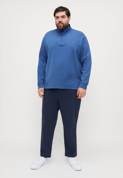 Blue fleece pullover with a half zipper and a logo on the chest, paired with navy pants and white sneakers. Soft texture, relaxed fit.
