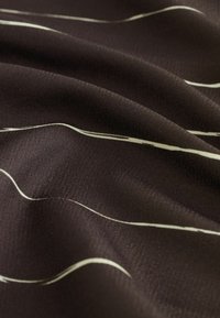 Tamno smeđi materijal s glatkom teksturom, ukrašen bijelim valovitim linijama koje stvaraju suptilan uzorak kroz cijeli materijal.