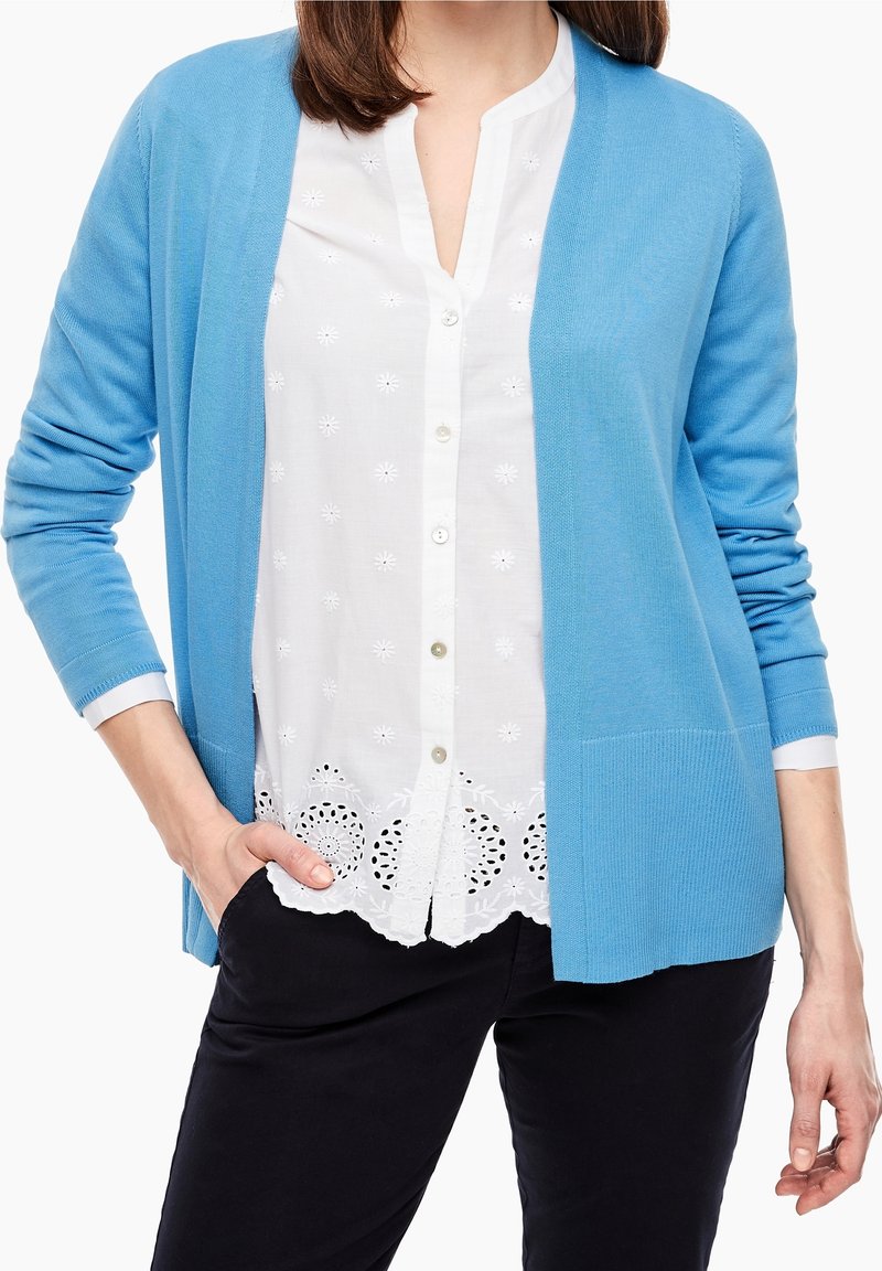 Světle modrý kardigan s dlouhými rukávy, otevřeným předním dílem, žebrovanou strukturou a bočními kapsami. Pod ním bílá vyšívaná blůza s knoflíkovou légou.
