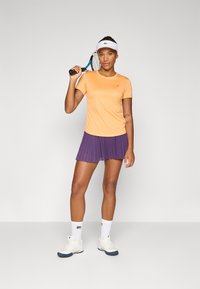 Person stands confidently with a tennis racket, wearing an orange shirt and purple pleated skirt, sporting white socks and shoes.