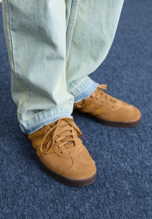 Personne portant un jean bleu clair et des baskets en daim marron debout sur un sol moquetté bleu.