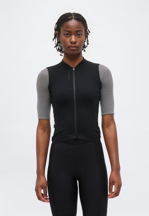 Mujer con el pelo trenzado, vistiendo un maillot de ciclismo negro ajustado con mangas cortas grises y pantalones cortos de ciclismo negros, de pie frente a un fondo blanco.