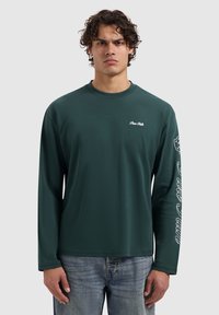 Donkergroene langemouwen t-shirt gemaakt van een gladde stof. Heeft witte tekst op de linkerborst en langs de rechtermouw. Casual pasvorm.