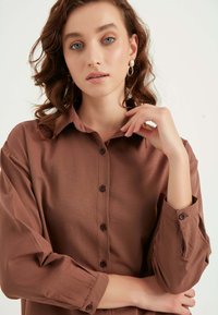 Camicia button-up marrone a maniche lunghe con una texture liscia. Presenta un colletto e polsini arrotolati con dettagli di bottoni.