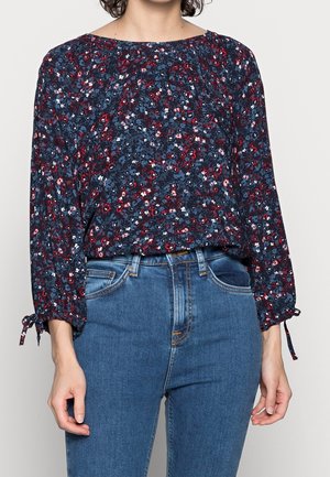 Femme portant un chemisier bleu marine à fleurs avec des manches longues nouées aux poignets, associé à un jean bleu taille haute.