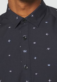 Camicetta nera con colletto, caratterizzata da un motivo geometrico in blu chiaro e bianco. Il tessuto sembra liscio e leggero.