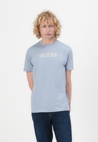 Vaaleansininen puuvillainen t-paita lyhyillä hihoilla, jossa on edessä valkoinen "GUESS"-logo ja perustamisvuosi painettuna. Rennon sovituksen malli.