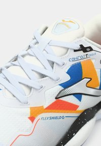 Balti kedu ar krāsainiem oranžiem, zilajiem un dzeltenajiem akcentiem, baltiem šņorēm un uzrakstiem "CONFORT Tongue" un "FLEXS HiELD" sānos.