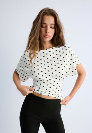 Young woman with long wavy hair wearing a white polka dot top and black pants adjusts her clothing against a plain background.