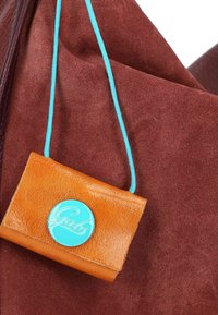 Braune Wildledertasche mit einer kleinen orangefarbenen Lederbörse, die mit einem türkisfarbenen Kordel verbunden ist, und einem blauen Knopf mit weißer Schrift.