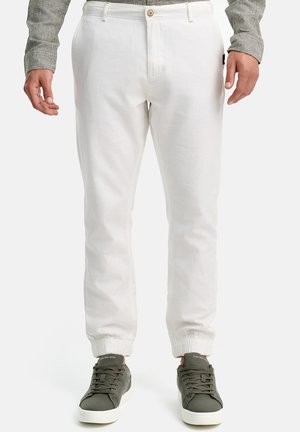 Mann trägt weiße Jogger-Hosen mit elastischen Bündchen und dunkelgrünen Sneakern, steht vor schlichtem Hintergrund.