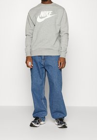 Ljusgrå sweatshirt med en stor vit Nike-logotyp, kombinerad med löst sittande blå jeans och svarta träningsskor med metalliska detaljer.