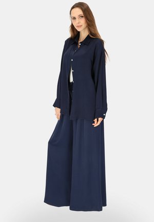 Kobieta w luźnej granatowej koszuli na guziki i szerokich spodniach, stojąca na jednolitym jasnym tle.