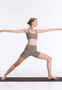 Femme en tenue de sport à carreaux effectue la posture de yoga Guerrier II sur un tapis noir devant un fond blanc uni.