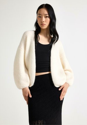 Donna con lunghi capelli neri che indossa un cardigan bianco peloso, un top nero corto e una gonna nera testurizzata su uno sfondo semplice.