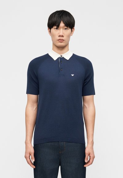 Homme portant un polo bleu marine à manches courtes avec col blanc et jean foncé, debout devant un fond gris clair uni.