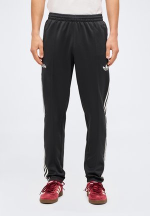 Osoba nosząca czarne dresowe spodnie Adidas z białymi paskami po bokach oraz czerwone buty sportowe Adidas, stojąca na jednolitym tle.
