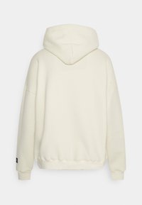 Bílá mikina s kapucí a volným střihem, která má měkkou textura a žebrované manžety. Zadní strana má hladký design bez grafik nebo akcentů.