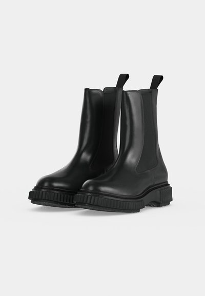 Paire de bottines Chelsea en cuir noir avec panneaux latéraux élastiques, languettes de traction et semelles épaisses en caoutchouc texturé sur un fond clair uni.