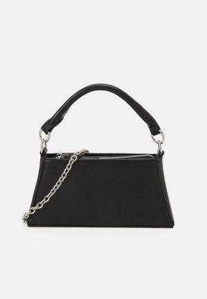 Bolso trapezoidal negro con asa de cuero redondeada, cadena plateada como correa y cierre superior con cremallera sobre un fondo liso y claro.