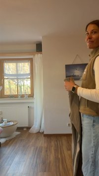 Brun fleeceväst, vit långärmad tröja, håller beige tyg. Ljust trägolv, vita gardiner och ett fönster med ett grått landskapsmotiv.