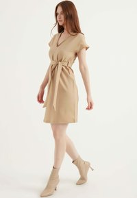 Beige Wickelkleid mit tiefem V-Ausschnitt, kurzen Ärmeln und gebundenem Taillenbereich. Glattes Material und Bleistiftrockform, kombiniert mit beigen Knöchelstiefeln.