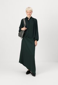 Tamsiai žalia, tekstūruota mygtukinė bliuska su raukšlėta apykakle, derinama su atitinkama asimetriška maksi sijonu; juodi mule batai ir pilka petnešų rankinė.