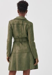 Manteau trench ceinturé vert olive, en matériau doux en suédine synthétique, avec un col et un ourlet évasé, agrémenté de coutures décoratives.