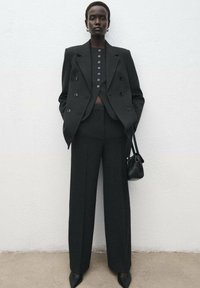 Blazer noir ajusté avec des boutons croisés, associé à un pantalon large taille haute. Les accessoires comprennent un sac à main noir et des talons pointus.