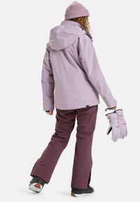Světle fialová nepromokavá bunda s kapucí, vrstvená přes tmavě vínové sněhové kalhoty. Světle fialové rukavice drženy v jedné ruce, černé boty s bílými podrážkami.