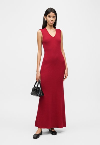 Κόκκινο, αμάνικο μακρύ φόρεμα με V-λαιμόκοψη, ριμπ υφή, εφαρμοστή σιλουέτα. Συνδυάζεται με μια μικρή μαύρη τσάντα και επίπεδα παπούτσια.