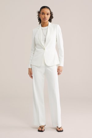 Frau mit lockigem Haar, die einen weißen, maßgeschneiderten Anzug, schwarze Sandalen und geschichtete silberne Halsketten vor einem schlichten beigen Hintergrund trägt.