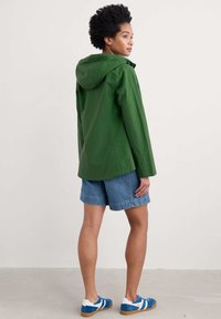 Zielona kurtka z kapturem wykonana z gładkiego materiału, posiadająca prosty design. W połączeniu z dżinsowymi szortami i niebieskimi trampkami z białymi akcentami.