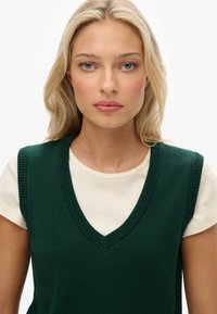 Groene gebreide vest met een diepe V-hals, gelaagd over een crèmekleurig T-shirt. Heeft ribgebreide randen en een gestructureerde stof, wat zorgt voor een casual uitstraling.