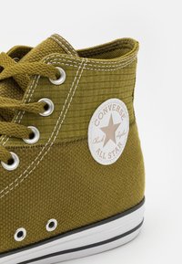 Sapatilha alta texturizada verde com ilhós de metal, costura branca e um emblema circular com o logótipo "Converse All Star".