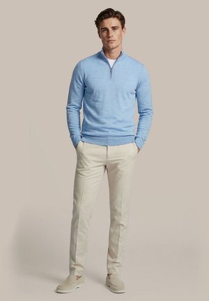 Man staat met handen in de zakken, draagt een lichtblauwe trui met kwart-ritssluiting, beige broek en beige instapschoenen tegen een effen achtergrond.