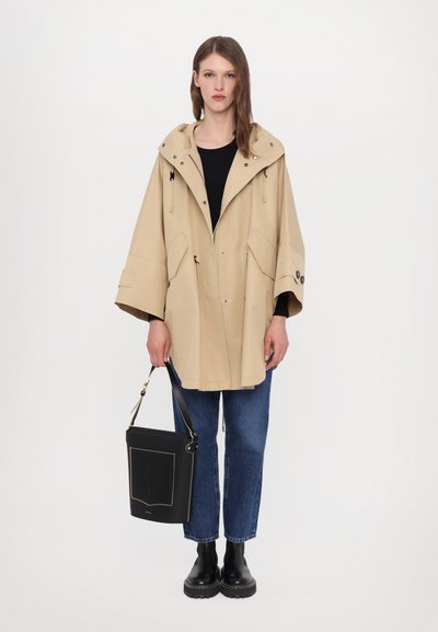 Beige oversize lietusmētelis ar lielām priekšējām kabatām, šņorējamām kapucēm un spiedpogu aizdari, kombinācijā ar tumšiem džinsiem un melniem masīviem zābakiem.