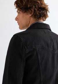 Ung mand med krøllet brunt hår iført en sort cowboyjakke over en ternet skjorte, kigger mod venstre mod en ensfarvet lys baggrund.