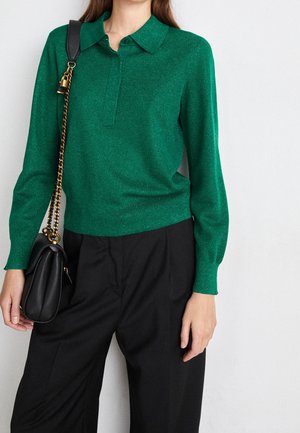 Femme portant un pull vert à manches longues avec col et un pantalon noir, portant un sac à main noir avec une bandoulière chaîne dorée sur l'épaule.