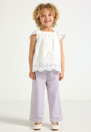 Smilende barn med krøllet hår iført hvid øjelygert top uden ærmer, lys lilla bukser med vide ben og hvide sko, stående på ensfarvet baggrund.