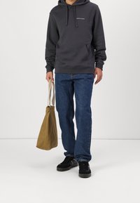 Felpa grigia scura con tasca frontale, jeans in denim blu e sneakers nere. Porta una borsa a tracolla marrone chiaro.