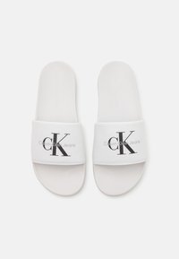 Hvite sandaler med glideskinn, som har en myk stoffstropp med svart "CK"-logo og "Calvin Klein Jeans"-tekst. Glatt såle med minimal tekstur.